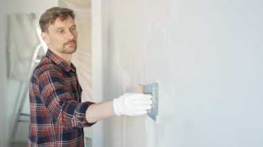 Eldiven takan ve elinde macun bıçağı tutan inşaat işçisi, beyaz bir duvara alçı yapıştırıyor, ev yenilemesi ve iyileştirme yapıyor.