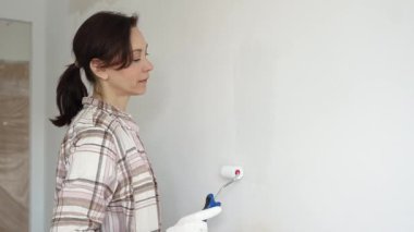 Koruyucu eldivenler giyen bir kadın, beyaz duvarı mavi bigudiyle boyuyor, iç mekanı güncelliyor ve yenileme projesinde odayı temizliyor, modern görünüme sahip..