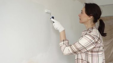 Koruyucu eldivenler giyen bir kadın, beyaz duvarı mavi bigudiyle boyuyor, iç mekanı güncelliyor ve yenileme projesinde odayı temizliyor, modern görünüme sahip..
