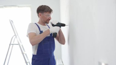 Güvenlik gözlüğü ve eldiven takan profesyonel tamirat ve montaj sırasında beyaz duvarda delik açmak için kablosuz matkap kullanıyor.