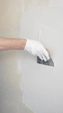 Eldiven giyen kimliği belirsiz kadın inşaat işçisi duvara sıva uygulamak için macun bıçağı kullanıyor. Ev yenileme projesi sırasında yüzeyi düzleştiriyor.