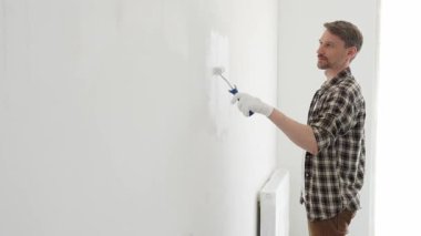 Profesyonel ressam beyaz duvarı silindirle düzleştiriyor, ev dekorasyonu sırasında koruyucu eldiven takıyor, iç mekanı yeni boyama tekniğiyle değiştiriyor.