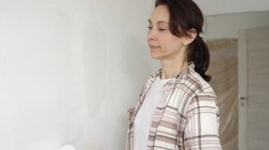 Koruyucu eldivenler giyen bir kadın, beyaz duvarı mavi bigudiyle boyuyor, iç mekanı güncelliyor ve yenileme projesinde odayı temizliyor, modern görünüme sahip..