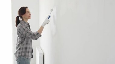 Eldiven giyen bir kadın boya silindiriyle beyaz bir duvar boyuyor, evini canlı bir ceketle değiştiriyor ve yenileme ve geliştirme sürecinin tadını çıkarıyor.