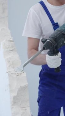 İnşaat işçisi, beyaz duvarı döner çekiç matkabıyla yıkıyor, mavi tulum ve koruyucu beyaz eldivenler giyiyor, yenileme projesi sırasında toz üretiyor, yakın çekim görüntüsü sağlıyor..