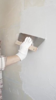 Eldiven giyen kimliği belirsiz kadın inşaat işçisi duvara sıva uygulamak için macun bıçağı kullanıyor. Ev yenileme projesi sırasında yüzeyi düzleştiriyor.