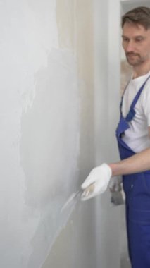Profesyonel inşaat işçisi mavi tulum ve beyaz eldiven giyerek, yenilenmiş, dikey görünümlü bir apartman dairesine beyaz bir duvar örmek için mala kullanıyor.