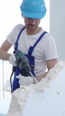 İnşaat işçisi beton duvarı döner çekiçle yıkıyor, koruyucu güvenlik giysileri giyiyor, yenileme projesi sırasında toz ve enkaz üretiyor. Dikey portre görünümü.
