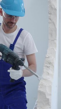 İnşaat işçisi beton duvarı döner çekiçle yıkıyor, koruyucu güvenlik giysileri giyiyor, yenileme projesi sırasında toz ve enkaz üretiyor. Dikey portre görünümü.