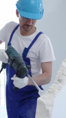 İnşaat işçisi beton duvarı döner çekiçle yıkıyor, koruyucu güvenlik giysileri giyiyor, yenileme projesi sırasında toz ve enkaz üretiyor. Dikey portre görünümü.