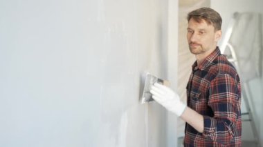 Eldiven takan ve elinde macun bıçağı tutan inşaat işçisi, beyaz bir duvara alçı yapıştırıyor, ev yenilemesi ve iyileştirme yapıyor.