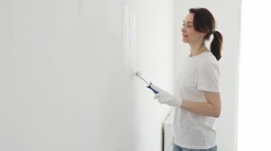 Beyaz eldiven giyen mutlu bir kadın boya silindirli beyaz bir duvar boyuyor. Ev yenileme sürecinin tadını çıkarıyor ve yeni bir görünüm yaratıyor.