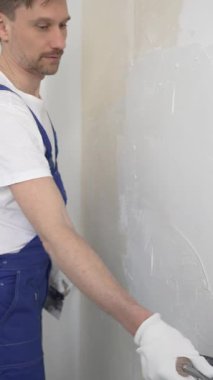 Profesyonel inşaat işçisi mavi tulum ve beyaz eldiven giyerek, yenilenmiş, dikey görünümlü bir apartman dairesine beyaz bir duvar örmek için mala kullanıyor.