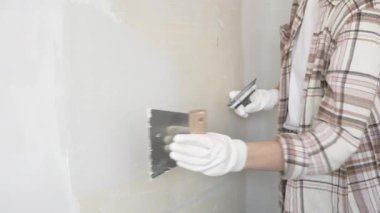 Eldiven giyen kimliği belirsiz kadın inşaat işçisi duvara sıva uygulamak için macun bıçağı kullanıyor. Ev yenileme projesi sırasında yüzeyi düzleştiriyor.