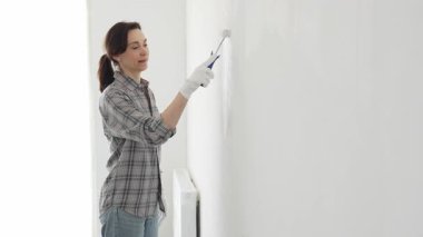 Eldiven giyen bir kadın boya silindiriyle beyaz bir duvar boyuyor, evini canlı bir ceketle değiştiriyor ve yenileme ve geliştirme sürecinin tadını çıkarıyor.