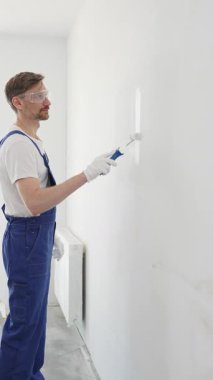 Beyaz eldivenler ve mavi tulumlar giyen profesyonel boyacı boş bir odada temiz beyaz bir duvar boyuyor, yenileme ve iç dekorasyona odaklanıyor.