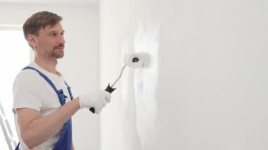 Mavi üniformalı erkek profesyonel ressam, iç dekorasyon projesi sırasında düzgün bir yüzey oluşturarak, duvarda dikkatlice beyaz boya sürüyor..