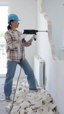 Kadın inşaat işçisi çekiç matkapla duvarı yıkıyor, koruyucu giysiler giyiyor, yenileme projesi sırasında moloz yığını oluşturuyor..