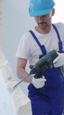İnşaat işçisi beton duvarı döner çekiçle yıkıyor, koruyucu güvenlik giysileri giyiyor, yenileme projesi sırasında toz ve enkaz üretiyor. Dikey portre görünümü.