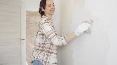 Kadın duvara macun bıçağıyla alçı yapıştırıyor, koruma için eldiven takıyor ve tadilat sırasında bile pürüzsüz ve sorunsuz bir şekilde bitmesini sağlıyor.