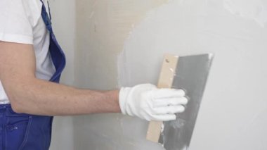 Bilinmeyen inşaat işçisi, beyaz duvar boyuyor, mavi tulum giyiyor ve apartman yenileme projesi sırasında koruyucu beyaz eldivenler giyiyor..