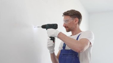Mavi tulumlu, güvenlik gözlüklü ve iş eldivenli bir inşaat işçisi kablosuz tornavida kullanarak duvara vida sokuyor.