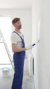 Beyaz eldivenler ve mavi tulumlar giyen profesyonel boyacı boş bir odada temiz beyaz bir duvar boyuyor, yenileme ve iç dekorasyona odaklanıyor.