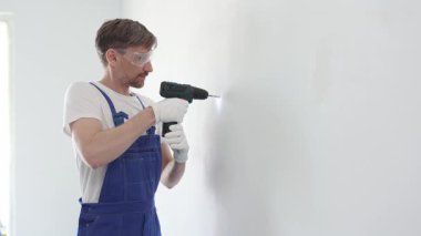 Güvenlik gözlüğü ve eldiven takan profesyonel tamirat ve montaj sırasında beyaz duvarda delik açmak için kablosuz matkap kullanıyor.