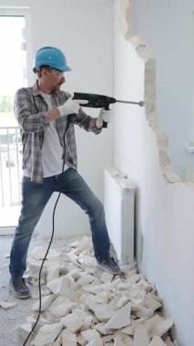 İnşaat işçisi, koruyucu teçhizatlı bir çekiç matkabıyla duvarı yıkıyor ve zemin yüzeyinde inşaat enkazı oluşturuyor. Yenileme konsepti.