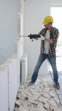 Koruyucu kask ve eldiven takan inşaat işçisi iç duvarı yıkmak için döner çekiç matkabı kullanıyor ve ev yenileme projesi sırasında yerde enkaz yığını oluşturuyor.