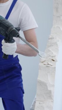 İnşaat işçisi, beyaz duvarı döner çekiç matkabıyla yıkıyor, mavi tulum ve koruyucu beyaz eldivenler giyiyor, yenileme projesi sırasında toz üretiyor, yakın çekim görüntüsü sağlıyor..