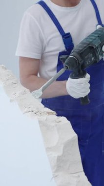 İnşaat işçisi, beyaz duvarı döner çekiç matkabıyla yıkıyor, mavi tulum ve koruyucu beyaz eldivenler giyiyor, yenileme projesi sırasında toz üretiyor, yakın çekim görüntüsü sağlıyor..