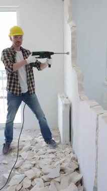 Koruyucu kask ve eldiven takan inşaat işçisi iç duvarı yıkmak için döner çekiç matkabı kullanıyor ve ev yenileme projesi sırasında yerde enkaz yığını oluşturuyor.