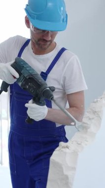 İnşaat işçisi beton duvarı döner çekiçle yıkıyor, koruyucu güvenlik giysileri giyiyor, yenileme projesi sırasında toz ve enkaz üretiyor. Dikey portre görünümü.