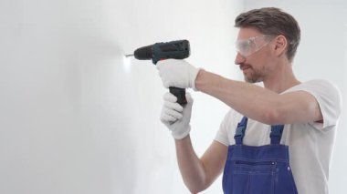 Profesyonel tamirci mavi üniformalı, güvenlik gözlüğü ve eldiven takıyor, tadilat ve montaj sırasında beyaz duvarda delik açmak için kablosuz matkap kullanıyor..
