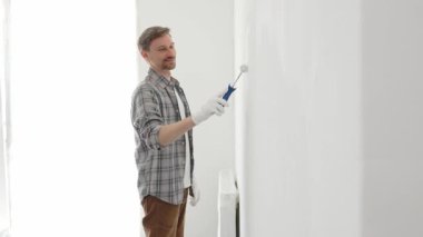 Profesyonel ressam iç duvarda beyaz boya yuvarlıyor, dikkatlice yüzeyini pürüzsüz bir şekilde kaplıyor, hatta ev yenileme projesi sırasında darbeler alıyor, kusursuz resim tekniğini vurguluyor.