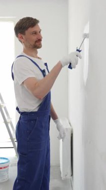 Beyaz eldivenler ve mavi tulumlar giyen profesyonel boyacı boş bir odada temiz beyaz bir duvar boyuyor, yenileme ve iç dekorasyona odaklanıyor.