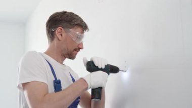 Güvenlik gözlüğü ve eldiven takan profesyonel tamirat ve montaj sırasında beyaz duvarda delik açmak için kablosuz matkap kullanıyor.