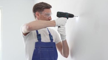Güvenlik gözlüğü ve eldiven takan profesyonel tamirat ve montaj sırasında beyaz duvarda delik açmak için kablosuz matkap kullanıyor.