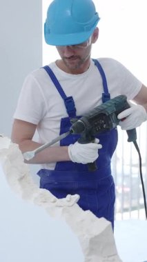 İnşaat işçisi beton duvarı döner çekiçle yıkıyor, koruyucu güvenlik giysileri giyiyor, yenileme projesi sırasında toz ve enkaz üretiyor. Dikey portre görünümü.