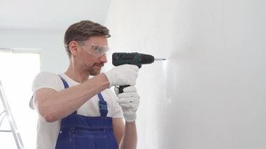 Mavi tulumlu, güvenlik gözlüklü ve iş eldivenli bir inşaat işçisi kablosuz tornavida kullanarak duvara vida sokuyor.