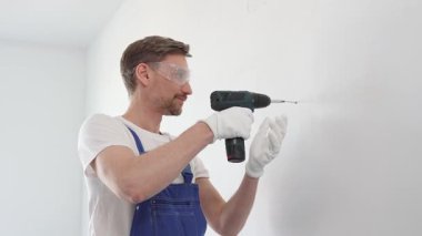 Mavi tulumlu, güvenlik gözlüklü ve iş eldivenli bir inşaat işçisi kablosuz tornavida kullanarak duvara vida sokuyor.