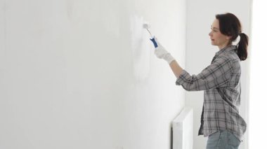 Eldiven giyen bir kadın boya silindiriyle beyaz bir duvar boyuyor, evini canlı bir ceketle değiştiriyor ve yenileme ve geliştirme sürecinin tadını çıkarıyor.