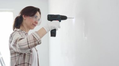 Güvenlik gözlüğü ve eldiven takan bir kadın, beyaz bir duvara kablosuz matkap kullanarak delik açıyor. Muhtemelen evindeki onarım ya da onarım projeleri için.