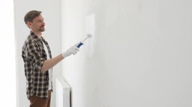 Profesyonel ressam beyaz duvarı silindirle düzleştiriyor, ev dekorasyonu sırasında koruyucu eldiven takıyor, iç mekanı yeni boyama tekniğiyle değiştiriyor.