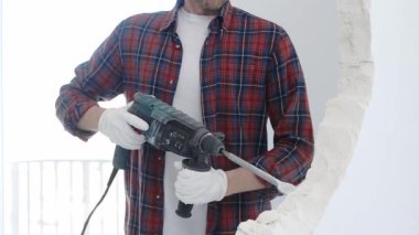 İnşaat işçisi, beyaz duvarı döner çekiç matkabıyla yıkıyor, kırmızı kareli gömlek ve koruyucu beyaz eldivenler giyiyor, yenileme projesi sırasında toz üretiyor, yakın çekim görüntüsü sağlıyor..