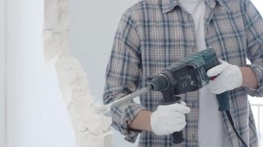 İnşaat işçisi, beyaz duvarı döner çekiçli matkapla yıkıyor, gri kareli gömlek ve koruyucu beyaz eldivenler giyiyor, yenileme projesi sırasında toz üretiyor, yakın plan görüntüsü var..
