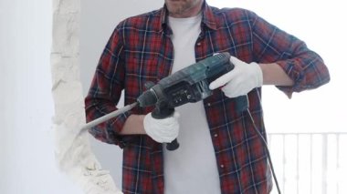 İnşaat işçisi, beyaz duvarı döner çekiç matkabıyla yıkıyor, kırmızı kareli gömlek ve koruyucu beyaz eldivenler giyiyor, yenileme projesi sırasında toz üretiyor, yakın çekim görüntüsü sağlıyor..