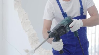 İnşaat işçisi, beyaz duvarı döner çekiç matkabıyla yıkıyor, mavi tulum ve koruyucu beyaz eldivenler giyiyor, yenileme projesi sırasında toz üretiyor, yakın çekim görüntüsü sağlıyor..