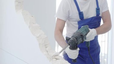 İnşaat işçisi, beyaz duvarı döner çekiç matkabıyla yıkıyor, mavi tulum ve koruyucu beyaz eldivenler giyiyor, yenileme projesi sırasında toz üretiyor, yakın çekim görüntüsü sağlıyor..